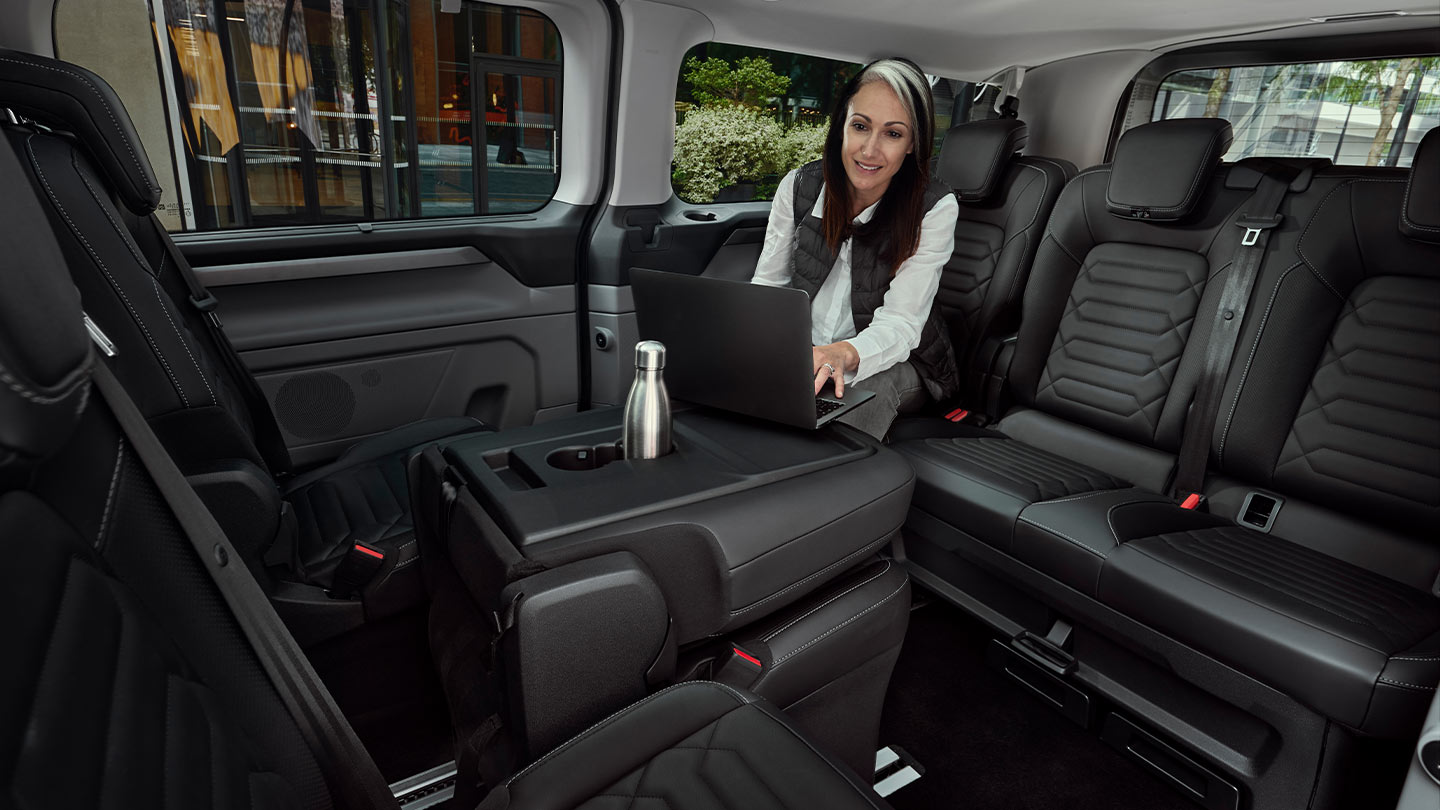 A woman sits at the table working on a laptop inside a Ford Tourneo Custom.