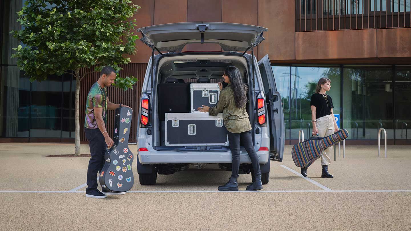 The rear of a silver Ford Tourneo Custom is open, with people loading musical equipment into the back.