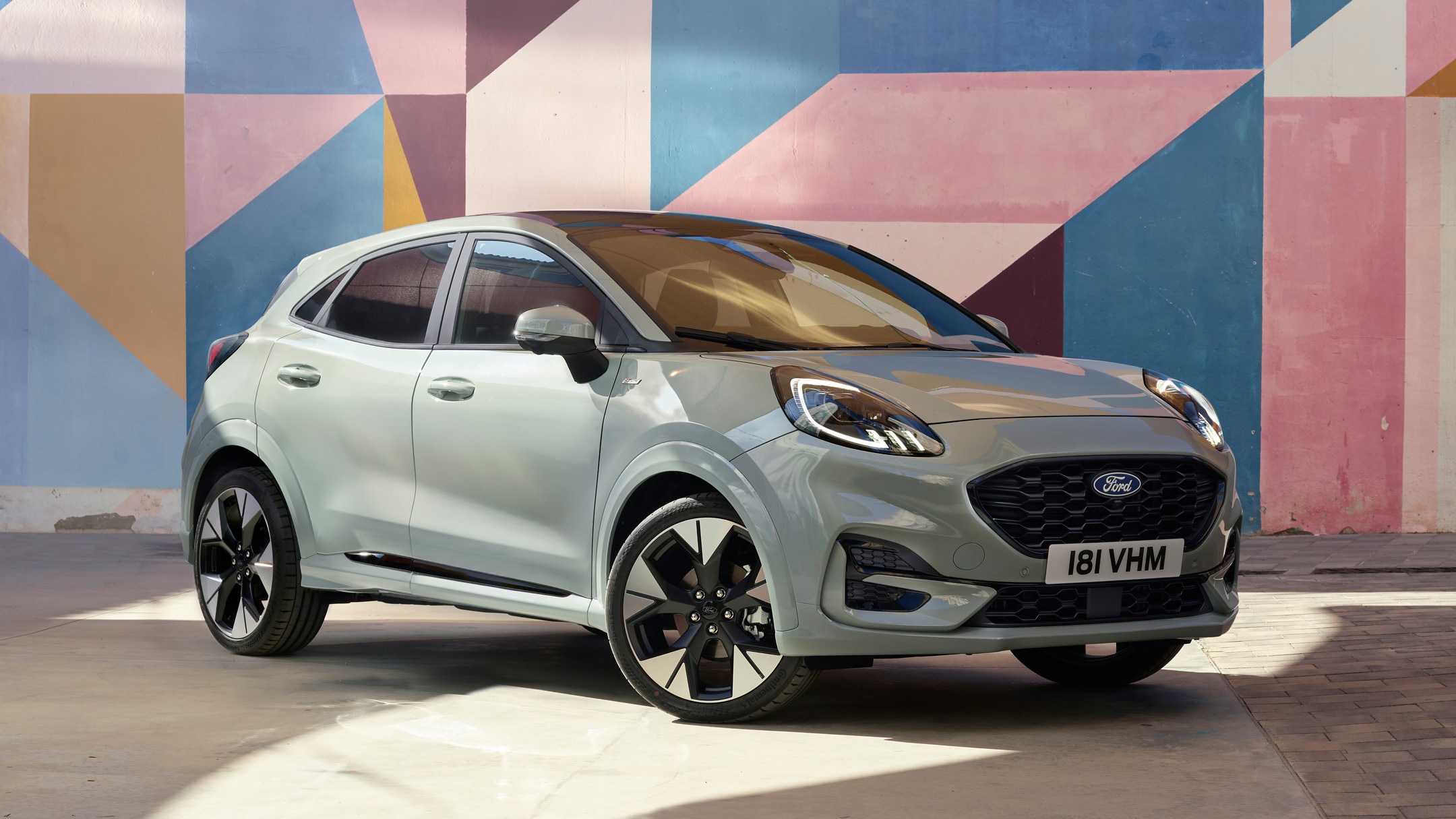 A Cactus Grey Ford Puma stands out against a backdrop of bold, geometric shapes, highlighting its modern design.